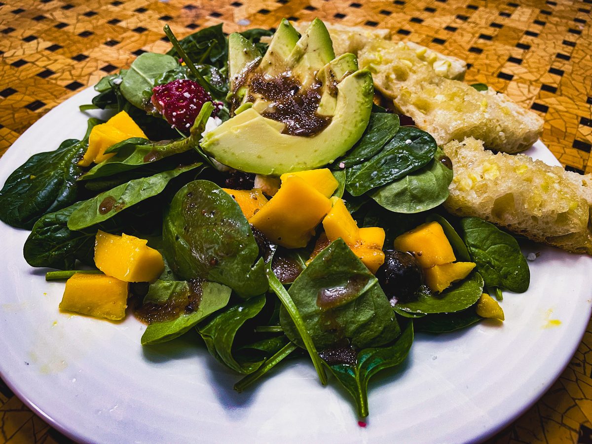 A plate of baby spinach salad with mango cubes, avocado slices, dark berries and a wedge of bread or cornbread.