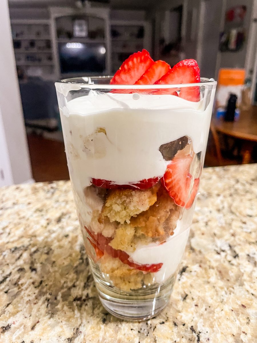 A strawberry shortcake parfait layered with whipped cream, cake crumbles and sliced strawberries in a tall pint glass on a granite