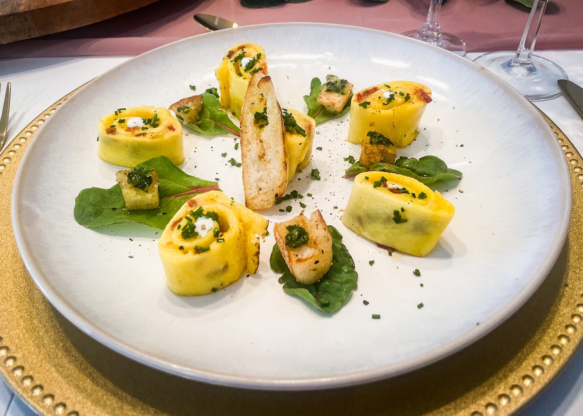 Plated appetizer of yellow pasta pinwheels with green herb sauce, small seared pale protein pieces on spinach leaves and a toasted