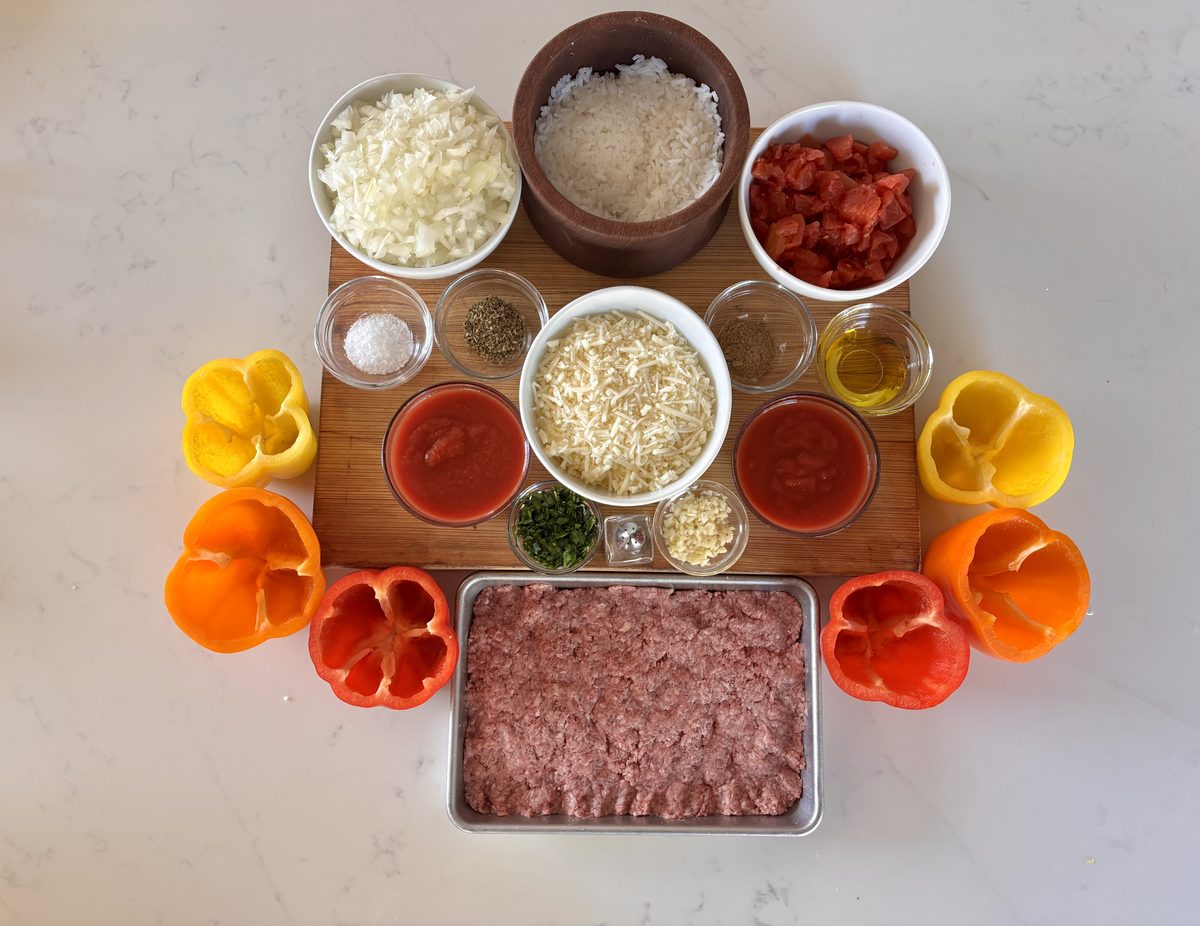 Overhead flat lay of stuffed-pepper prep on a marble counter: hollowed red, yellow and orange bell peppers ringing a wood board wi