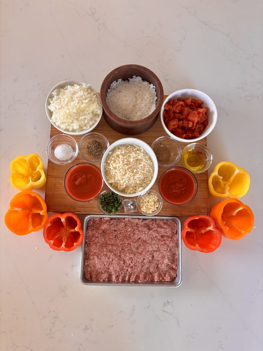 Same overhead ingredient flat lay in a tighter vertical crop: hollowed bell peppers around a board of rice, shredded cheese, diced