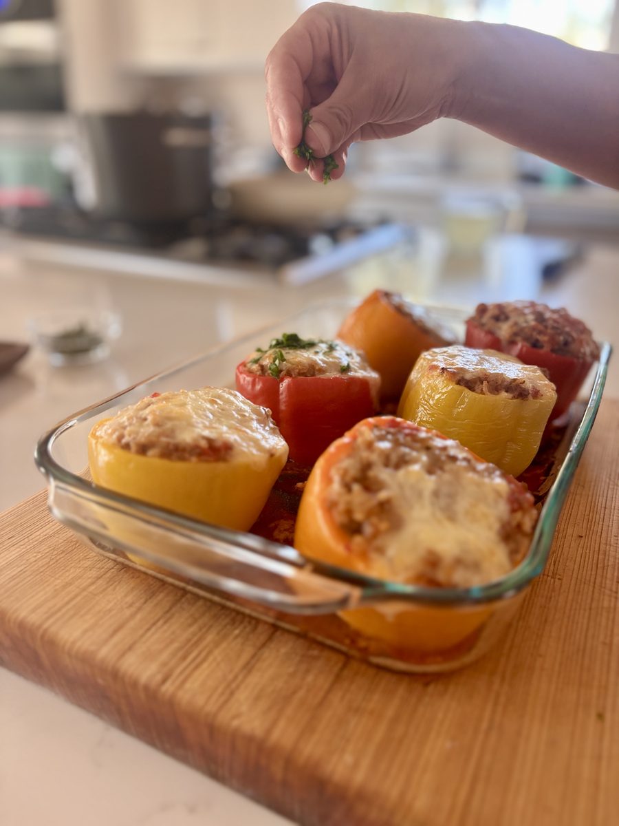 Baked stuffed bell peppers with melted cheese in a glass baking dish on a wooden board, a hand sprinkling chopped herbs from above