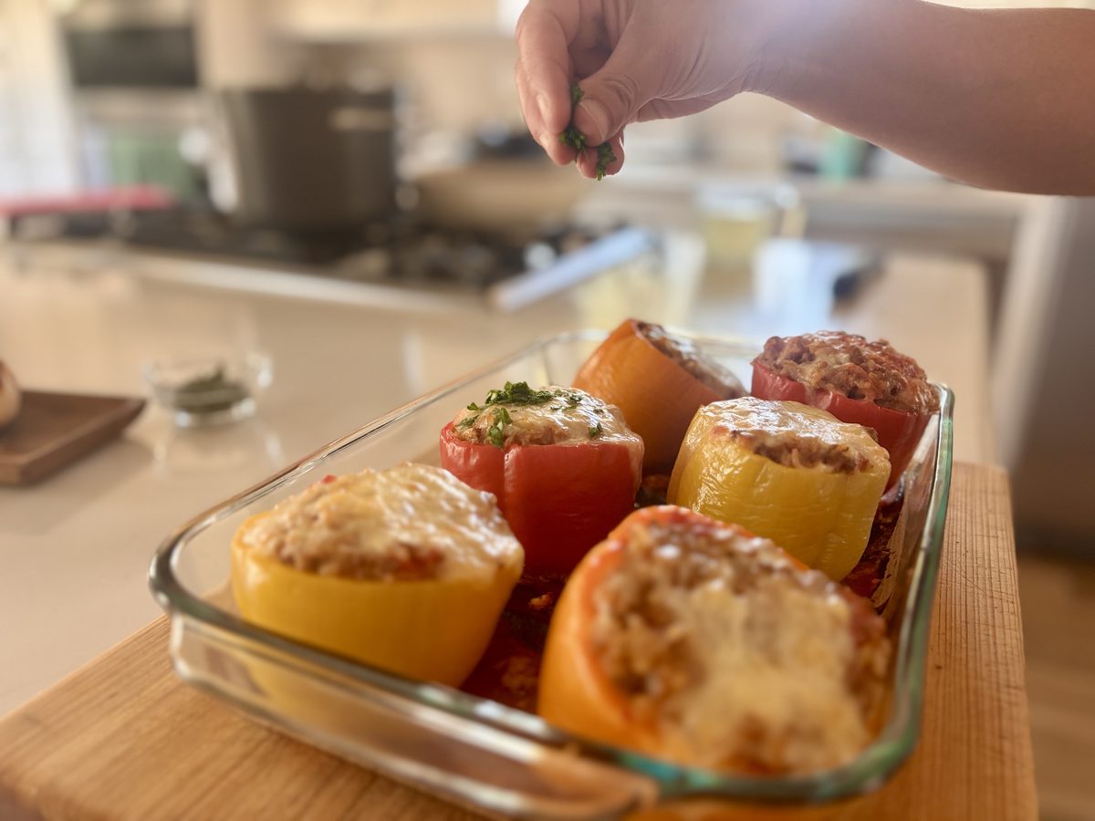 Closer view of baked cheese-topped stuffed peppers in a glass dish on a wood board, with a hand scattering fresh herbs over them.