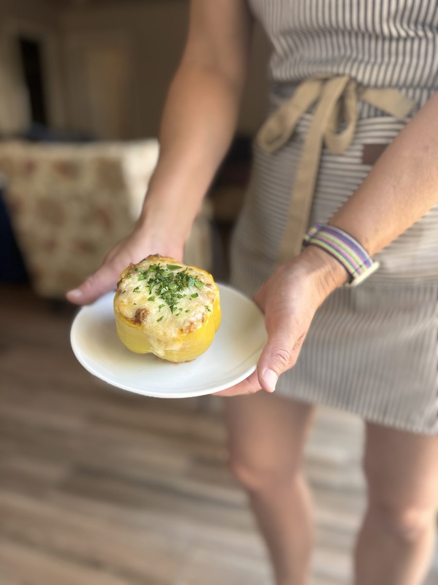 A woman in a striped apron holding a small white plate with one cheese-and-herb topped stuffed yellow pepper, standing on a wood f