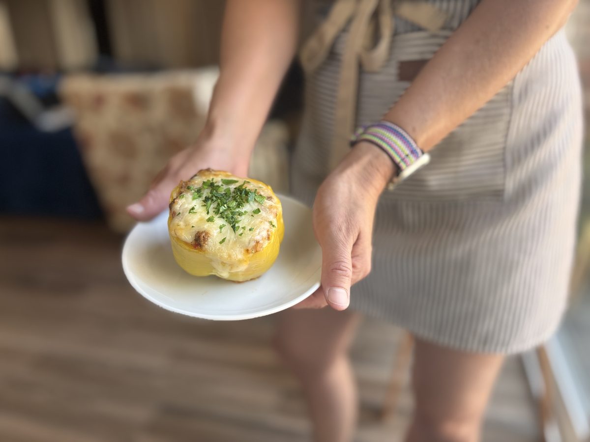 A woman in a striped dress holding out a small white plate with a whole baked yellow bell pepper stuffed with a creamy filling and