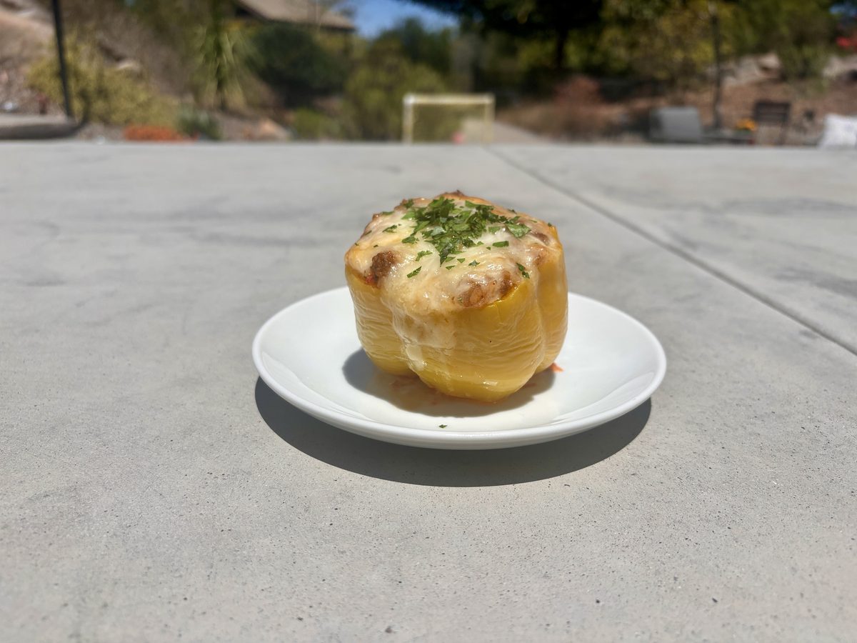A baked yellow stuffed pepper with browned cheesy filling and parsley on a small white plate set on an outdoor concrete table in b