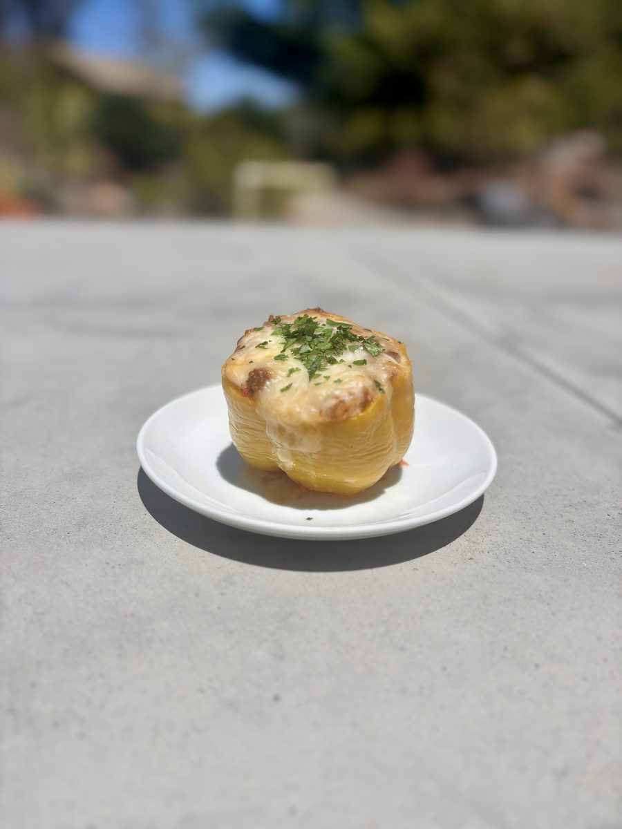 Distant shot of the same baked stuffed yellow pepper on a white plate on a sunlit concrete outdoor counter, subject small in frame