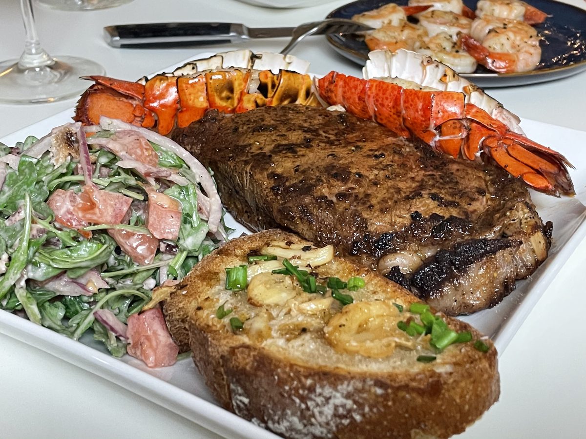 White platter of grilled steak with a split lobster tail, creamy arugula-tomato salad and a slice of garlic cheese toast, with a d