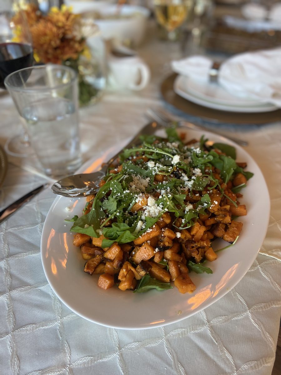 A white bowl-plate of roasted sweet potato cubes with arugula and crumbled feta on a formally set table with glassware and folded
