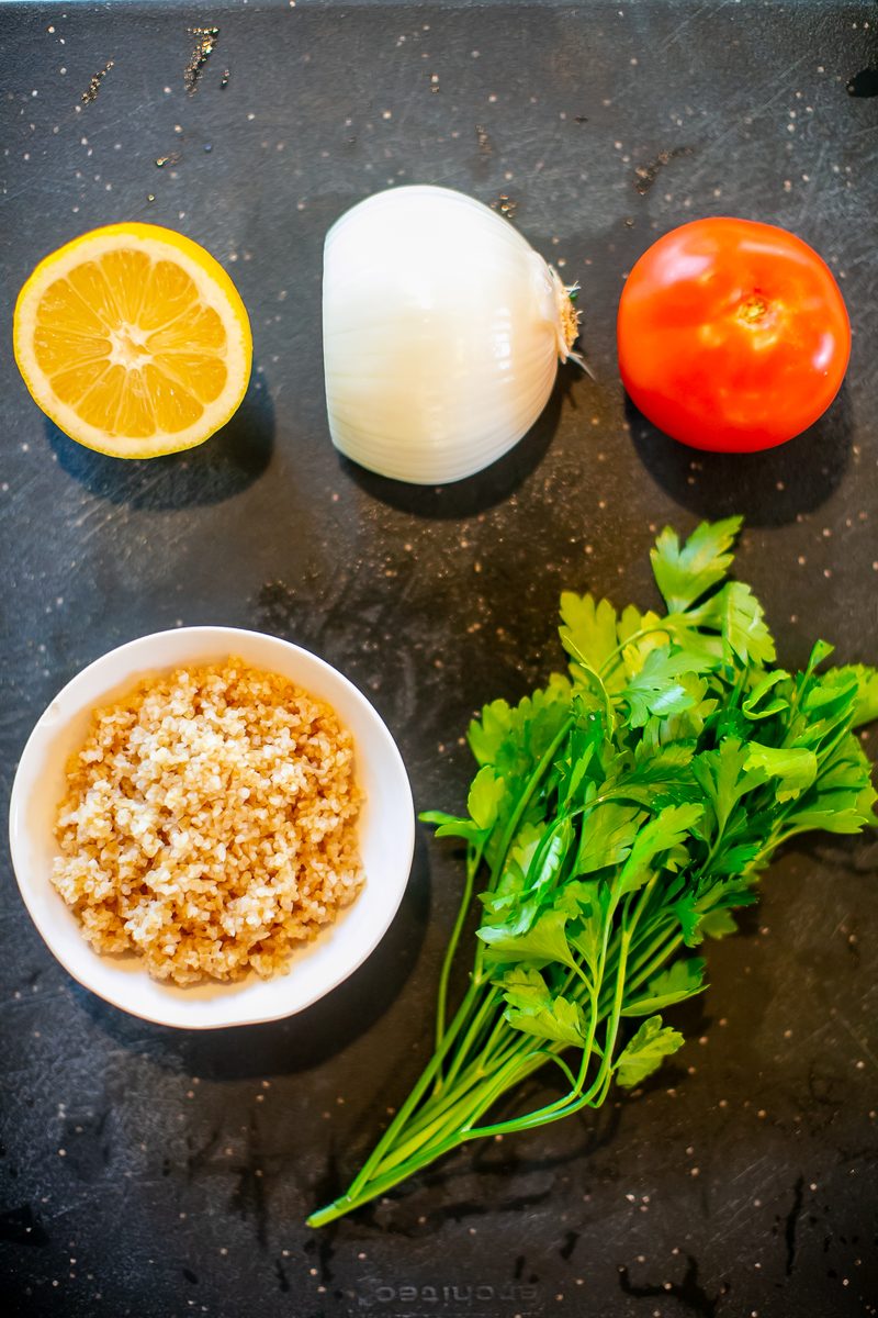 Overhead of tabouli ingredients on a dark slate board: half a lemon, a peeled onion, a tomato, a white bowl of soaked bulgur and a