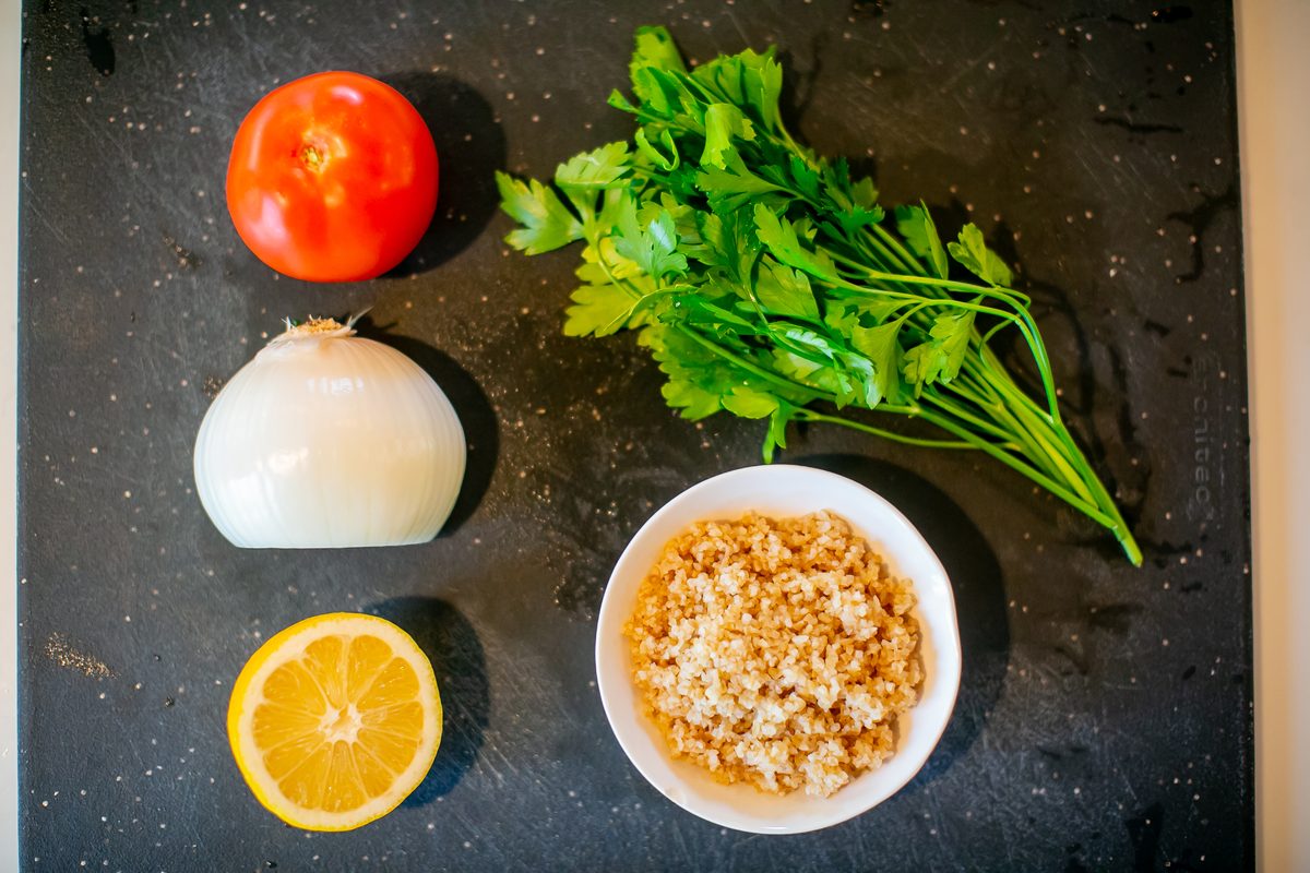 Angled overhead of the same tabouli ingredients on a dark slate board - tomato, onion, lemon half, parsley bunch and a white bowl