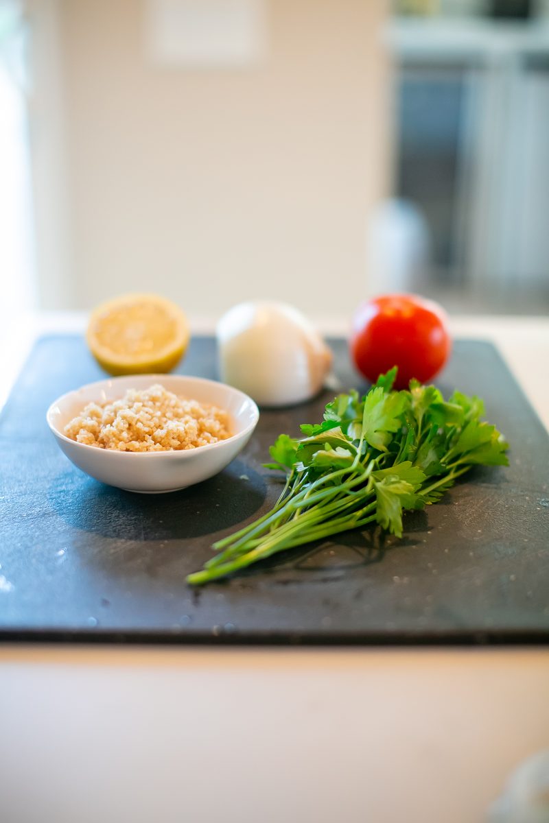 Wide, dim shot of the slate board of tabouli ingredients (bulgur bowl, lemon, onion, tomato, parsley) small in the frame on a kitc