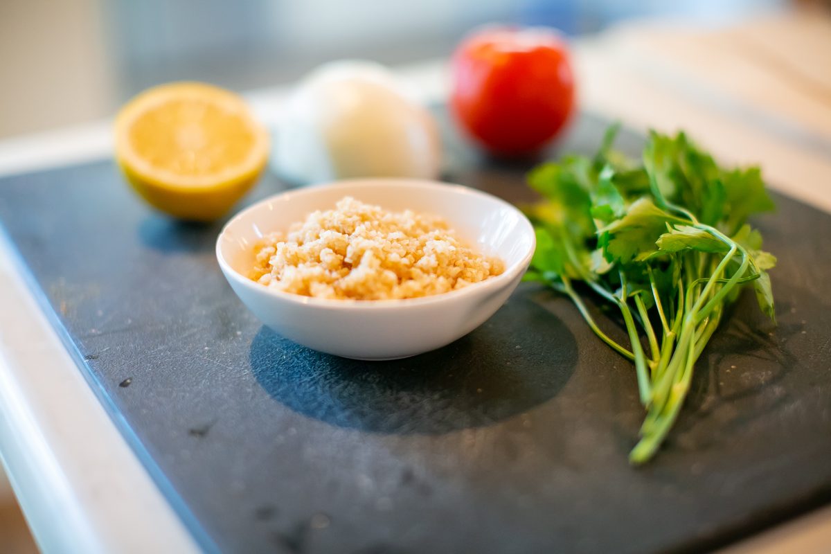 White bowl of soaked bulgur on a dark slate board with a lemon half, onion, tomato and parsley around it, soft daylight on a marbl