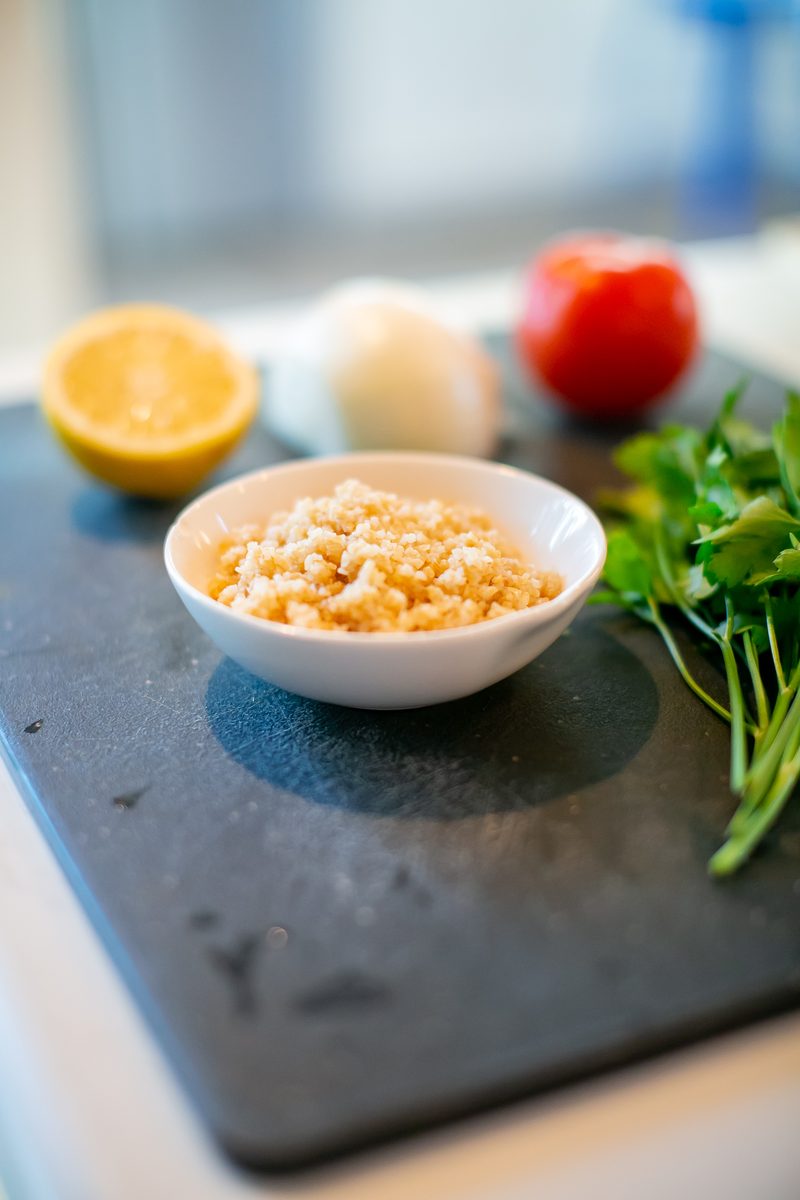 Tighter frame of the white bowl of bulgur on the slate board with lemon, onion, tomato and parsley alongside on a counter.