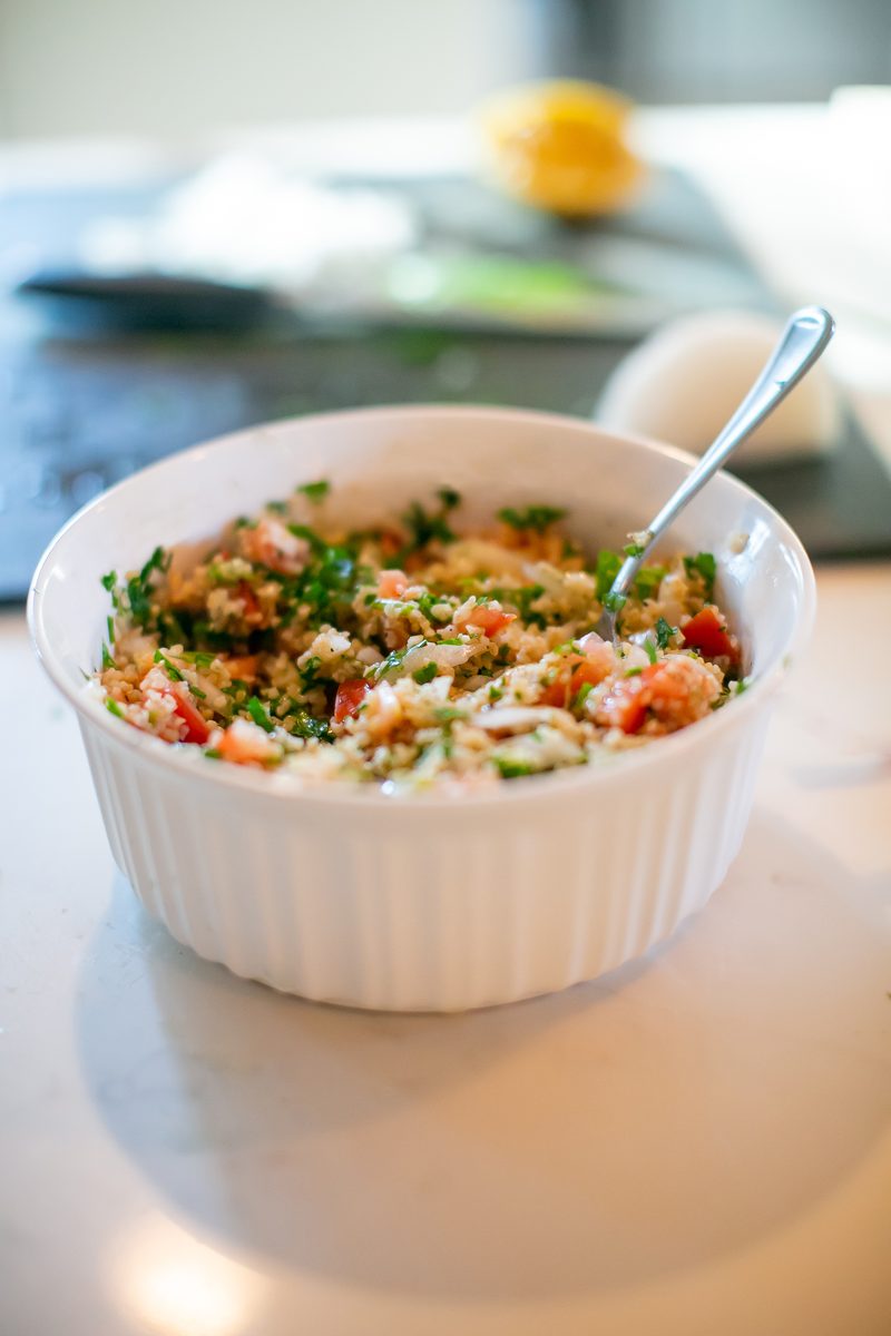 Finished tabouli salad of bulgur, chopped parsley and tomato in a white fluted bowl with a spoon standing in it, on a counter with