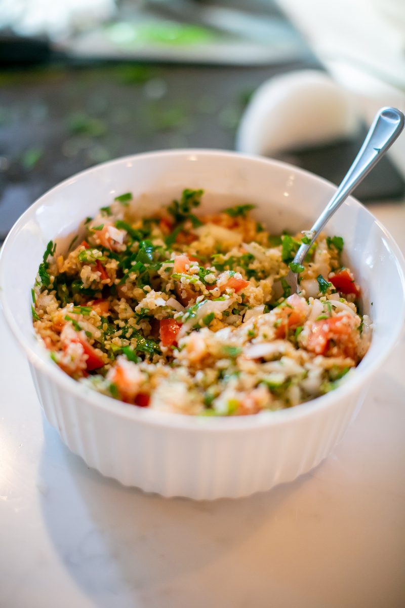 Close overhead of finished tabouli - bulgur with chopped parsley, tomato and onion - in a white ribbed bowl with a spoon.