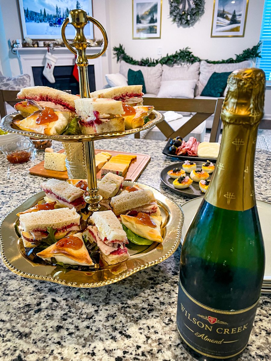 A two-tier gold serving stand of small crustless tea sandwiches and pastries beside a bottle of sparkling wine in a decorated room