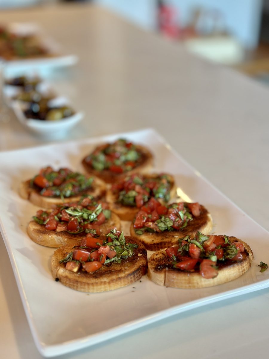 Nearly identical soft-focus shot of tomato-basil bruschetta rounds on a white tray on a kitchen counter with blurred bowls behind.