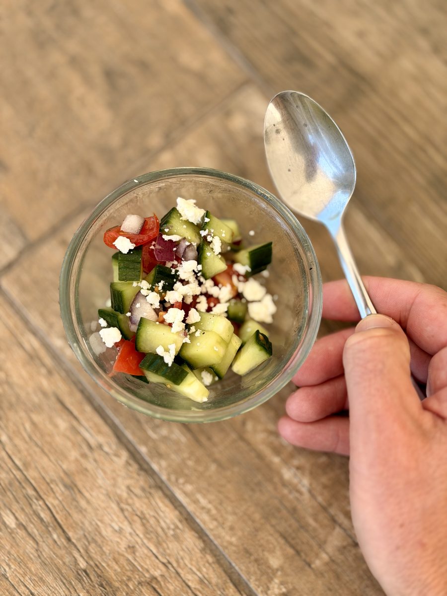 Overhead phone-style shot of a small glass bowl of chopped cucumber, tomato and crumbled feta on a wood plank floor, with a hand h