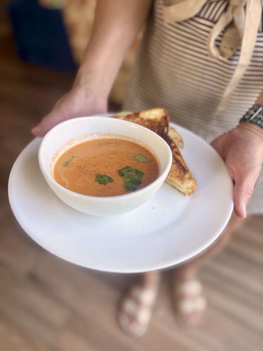 A woman holds a white plate with a bowl of creamy tomato soup garnished with basil and a half grilled cheese sandwich beside it.