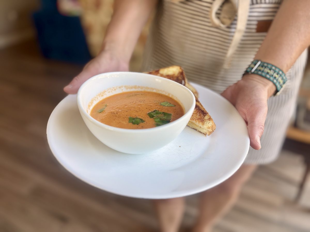 Closer view of hands holding the plated tomato soup bowl with basil garnish and a half grilled cheese sandwich, wood floor blurred