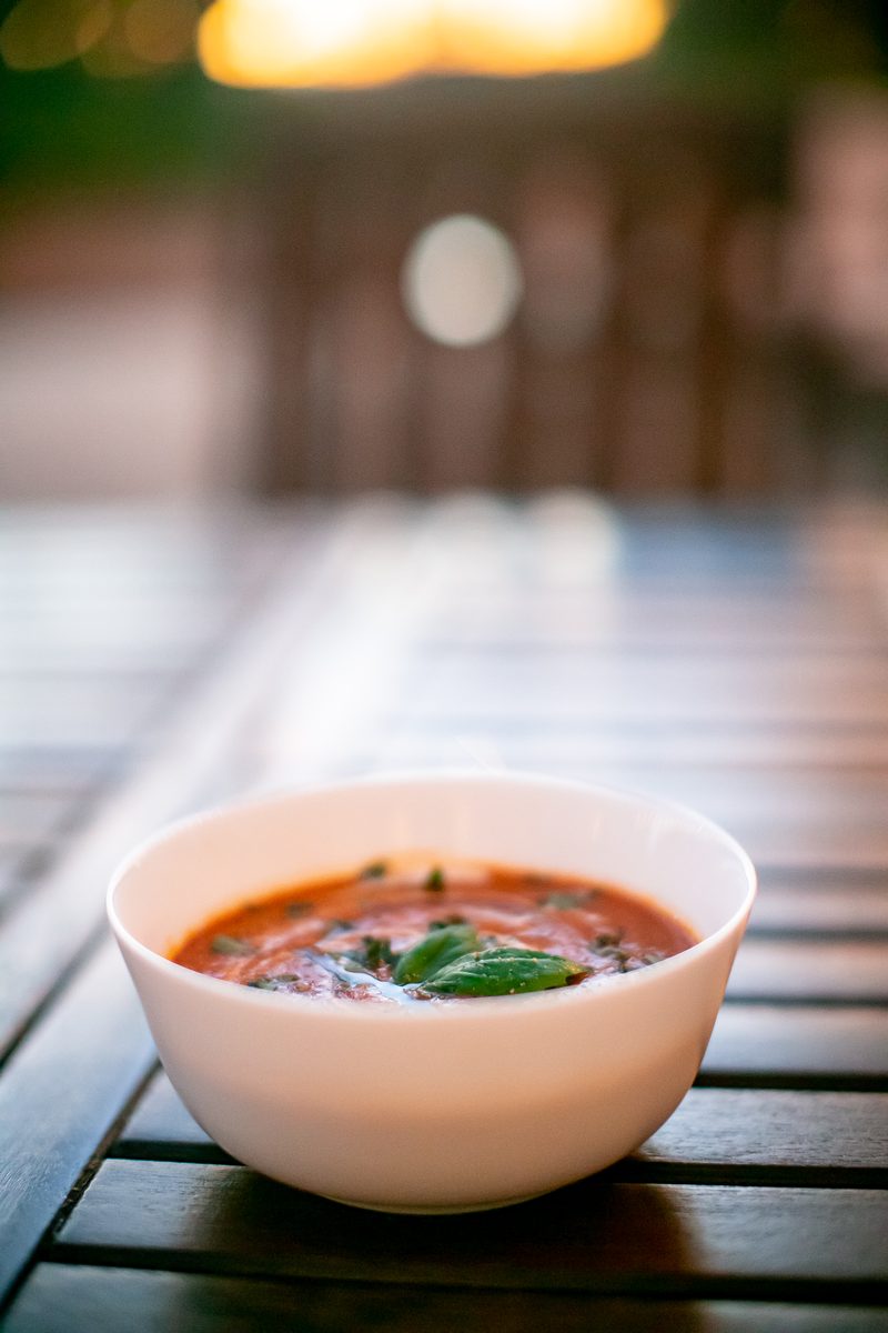 White bowl of creamy tomato soup garnished with basil on a slatted wooden outdoor table at dusk, warm string-light bokeh behind.