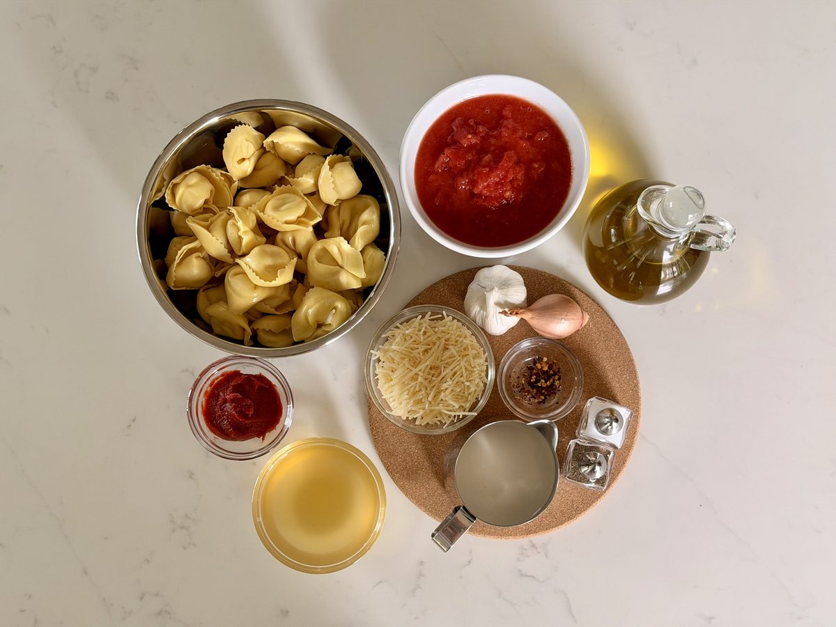 Overhead flat lay on marble of a metal bowl of dried tortellini, a white bowl of marinara, an oil cruet, grated parmesan, tomato p