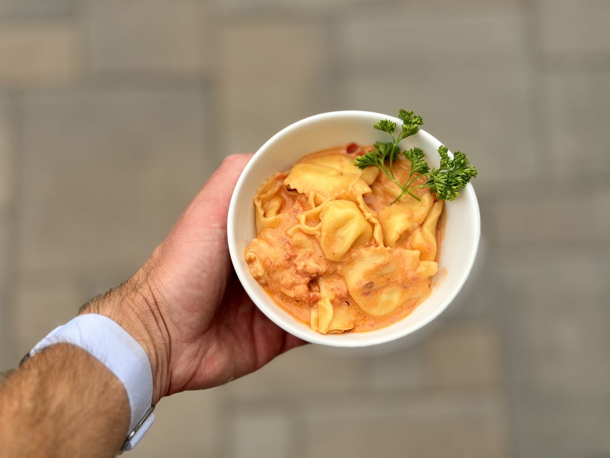 A hand holds a small white bowl of cheese tortellini in creamy tomato sauce garnished with a curly parsley sprig, stone wall blurr