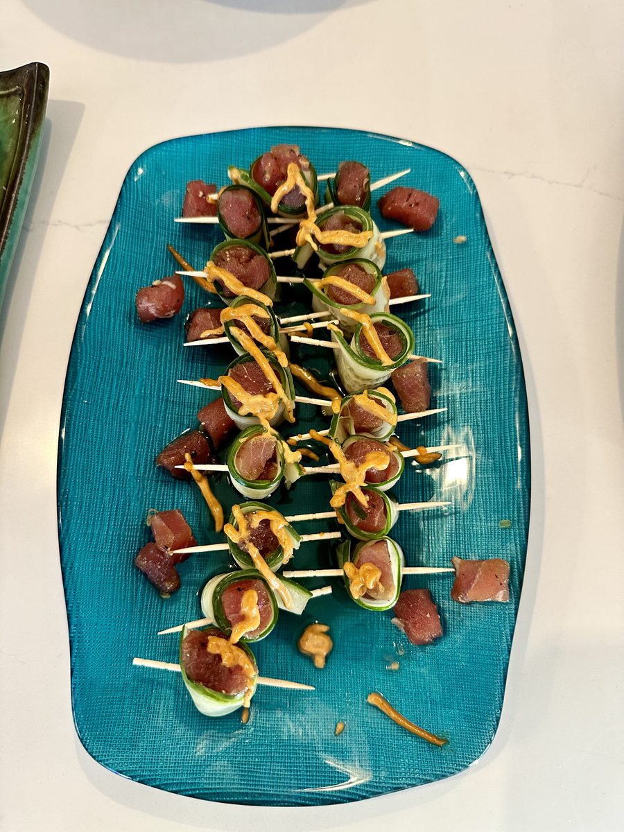 Toothpick skewers of raw tuna cubes wrapped in cucumber ribbons, drizzled with orange spicy mayo, arranged on a teal rectangular p