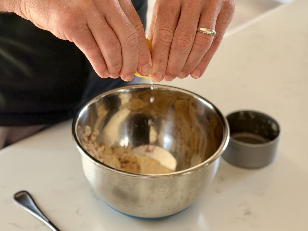 A man's hands squeeze a lemon wedge into a stainless mixing bowl holding a beige flaked salad mixture on a white counter; contents