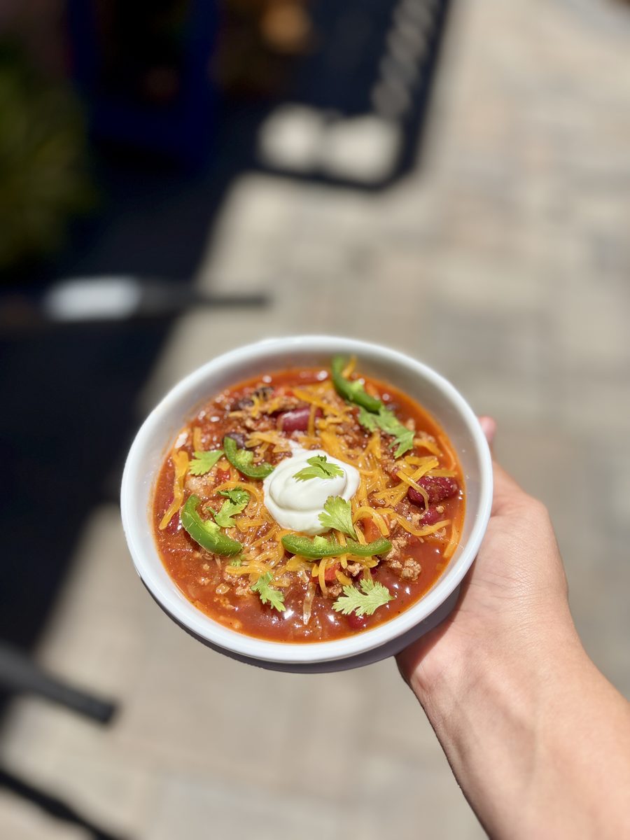 A hand holds a white bowl of chili with ground meat and kidney beans topped with shredded cheese, sour cream, jalapeno slices and