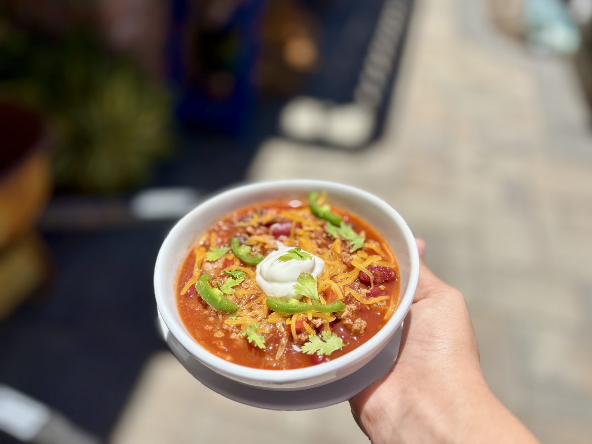 Close hand-held white bowl of chili loaded with ground meat, beans, shredded cheddar, a dollop of sour cream, jalapeno and cilantr