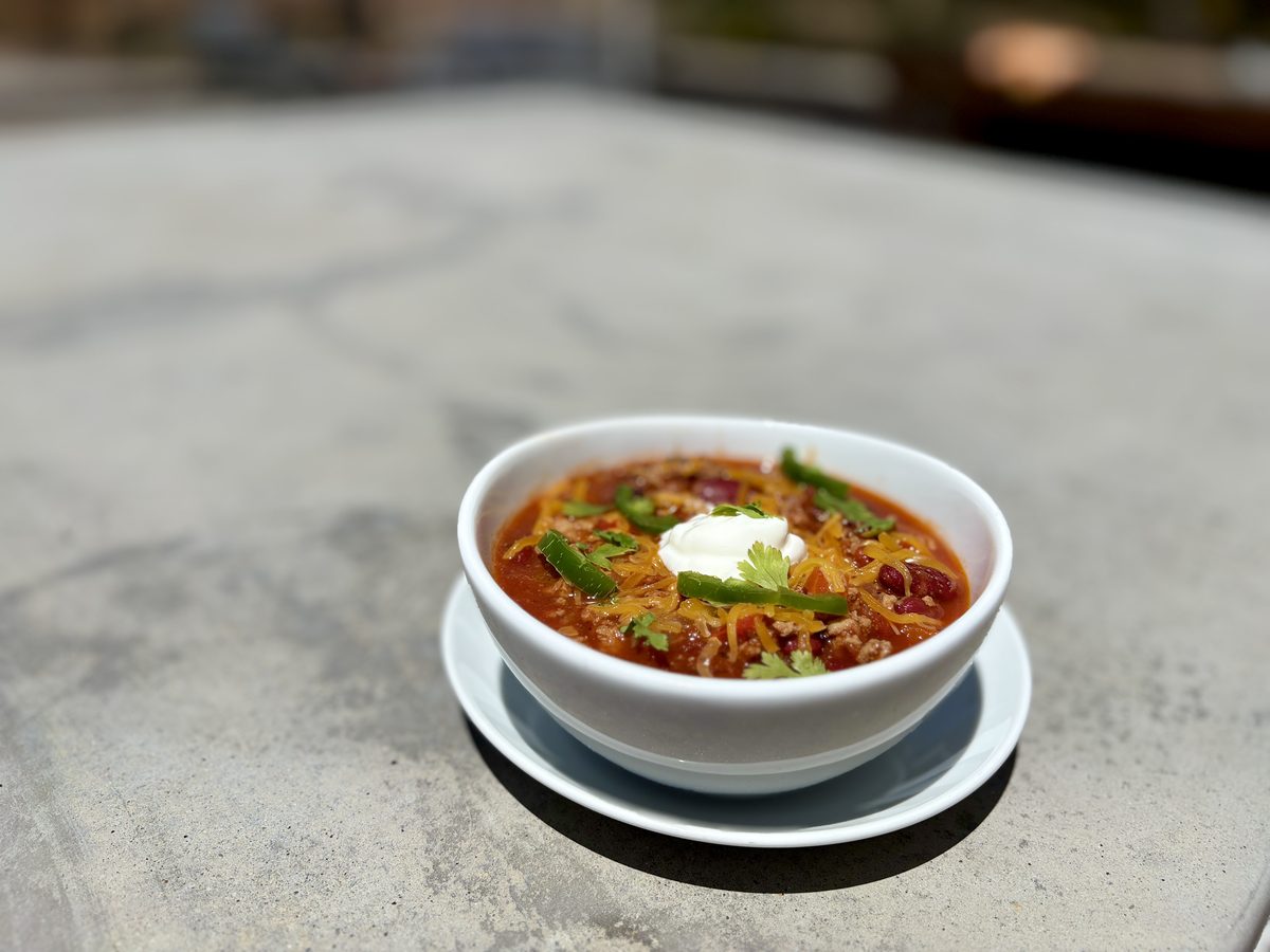 A white bowl on a saucer holding thick red chili with beans, shredded orange cheese, green pepper strips and a dollop of sour crea