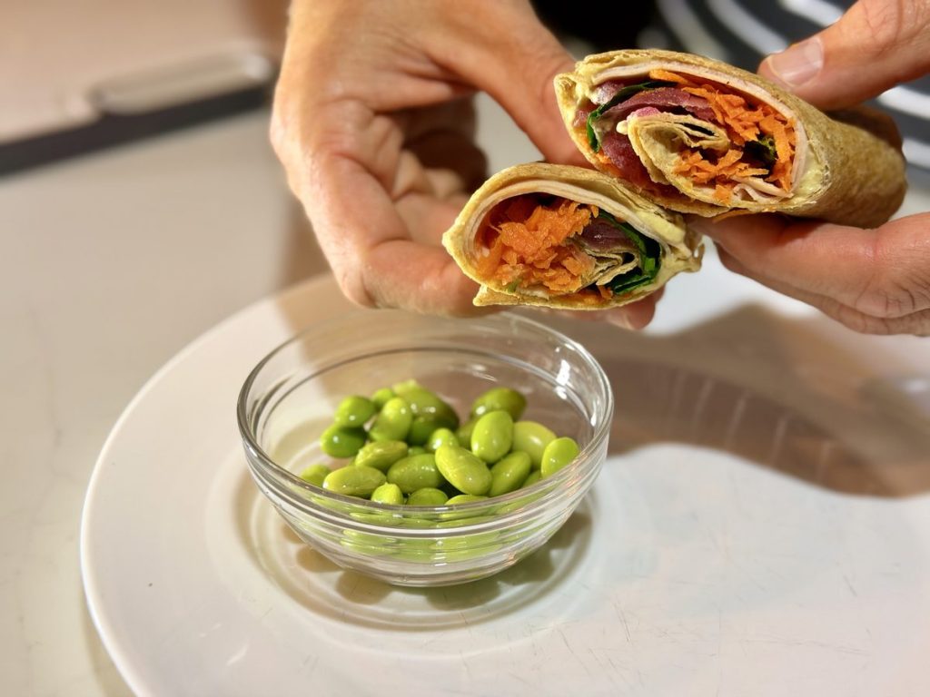 Two hands holding up the cut halves of a finished wrap showing filling of shredded carrot, red onion, spinach and pale sliced deli turkey, above a plate with a 