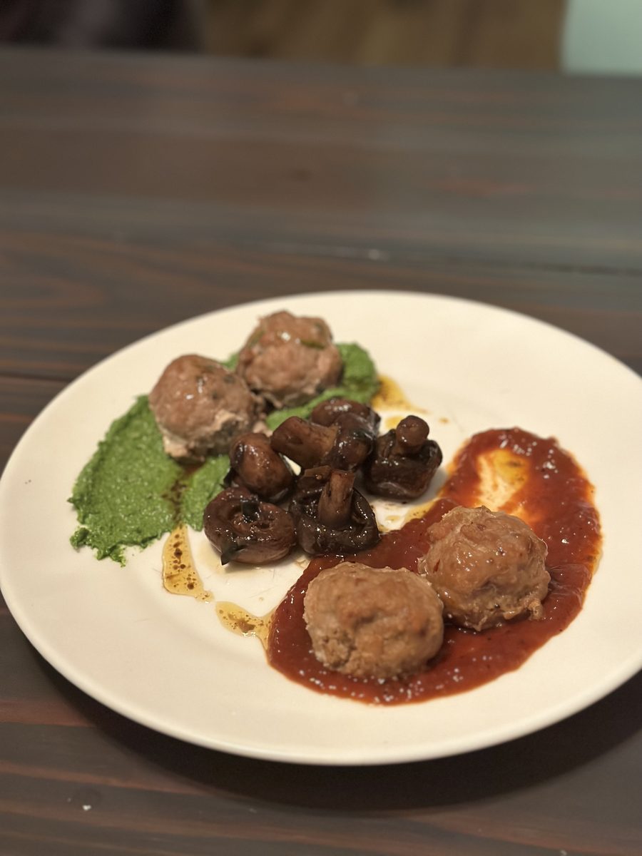 White plate of turkey meatballs with roasted mushrooms, a green pesto smear and a pool of marinara, on a dark wood table in dim li