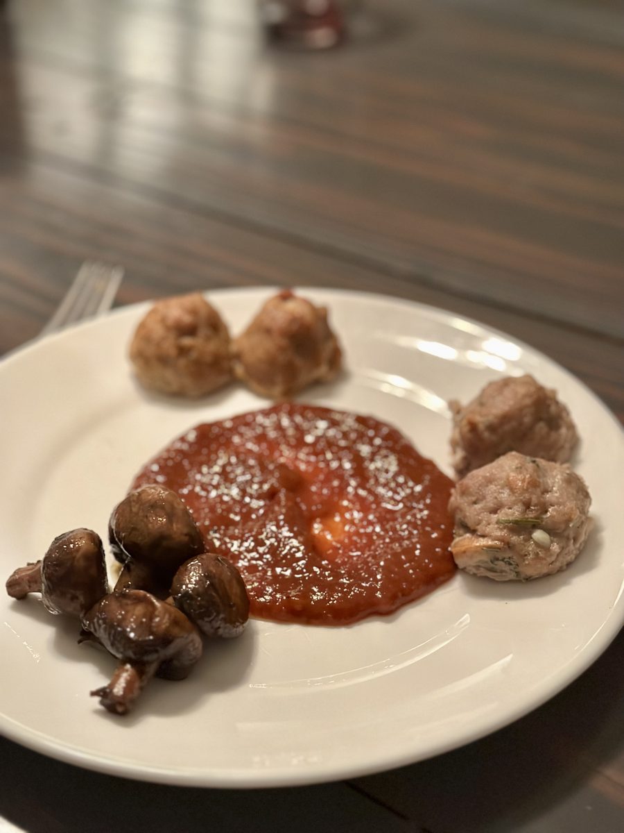 Overhead white plate with a few turkey meatballs, roasted mushrooms and a pool of marinara dusted with parmesan, on a dark wood ta