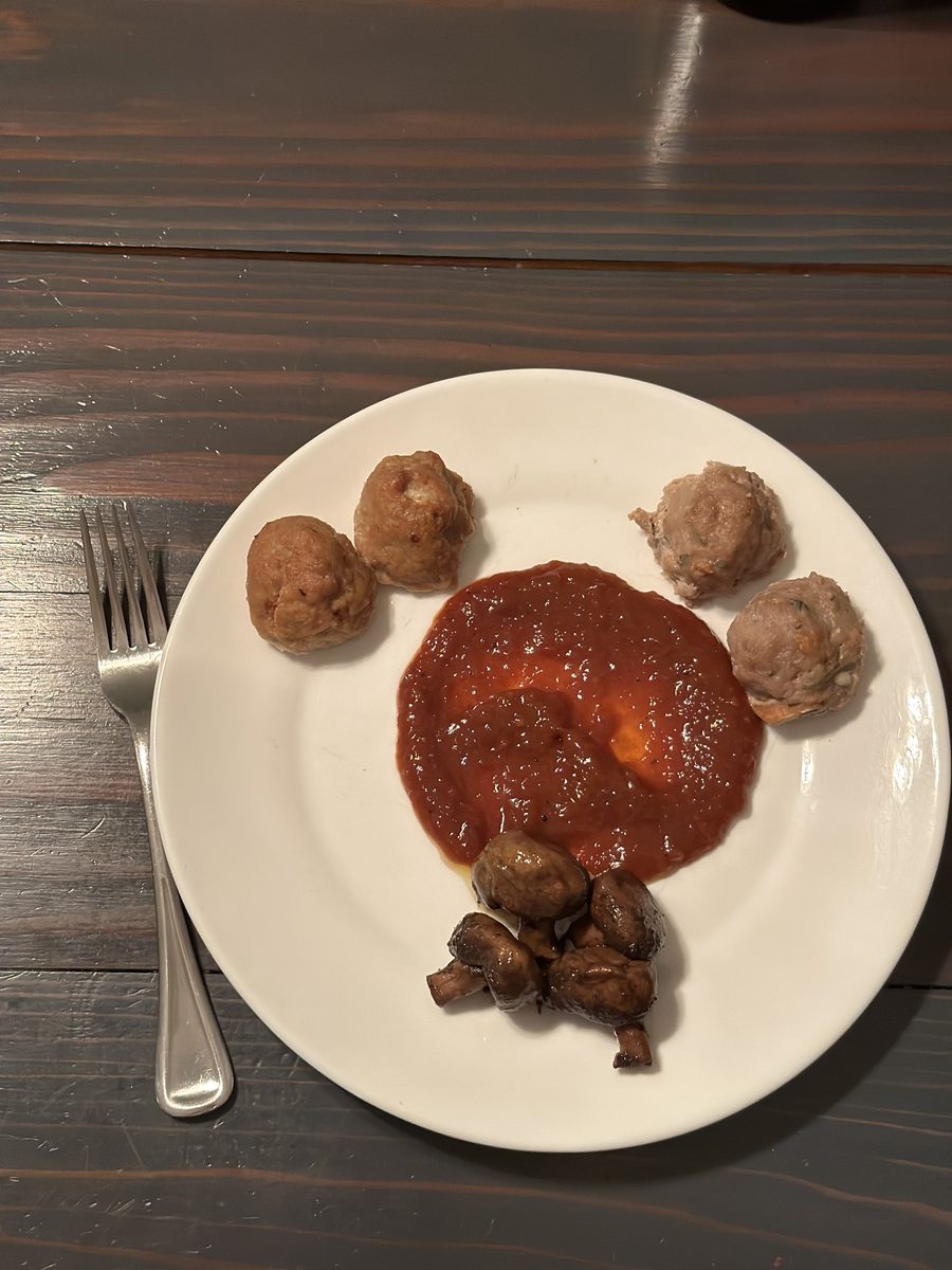 White plate of turkey meatballs, roasted mushrooms and marinara with a fork beside it on a dark wood table, dim sparse plating.