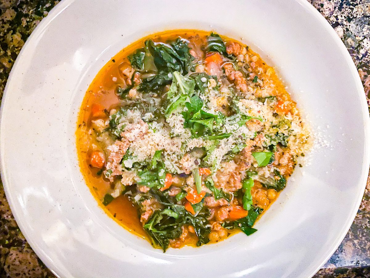 White bowl of Tuscan soup with crumbled sausage, white beans, carrot and greens under a heavy dusting of grated parmesan, on a gra