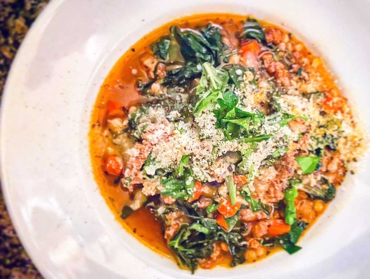 White bowl of brothy sausage and kale soup with beans and diced tomato topped with grated parmesan, on a speckled granite counter.