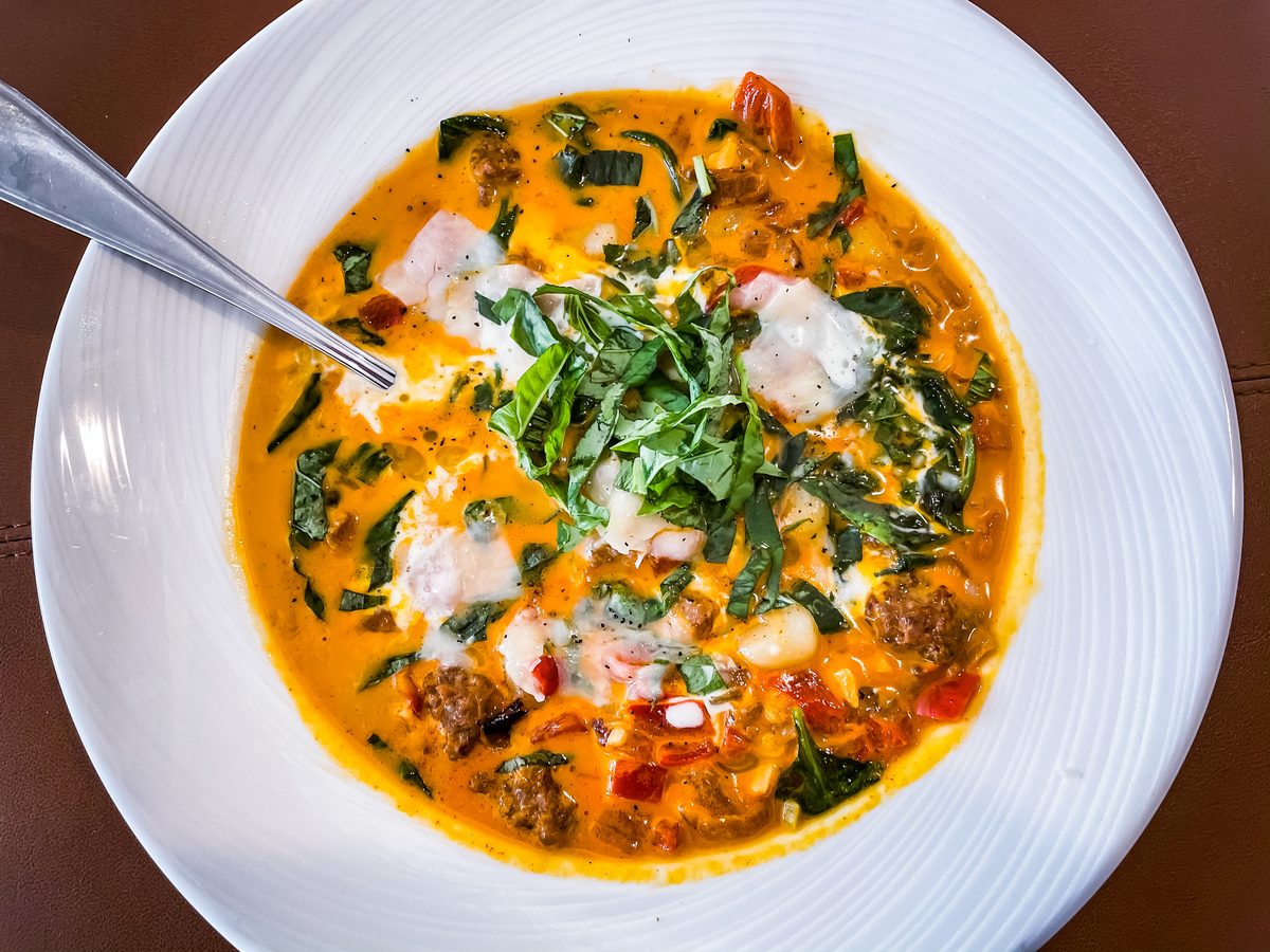 White bowl of creamy orange tomato soup with crumbled sausage, pieces of shrimp and shredded greens, spoon resting in the bowl on
