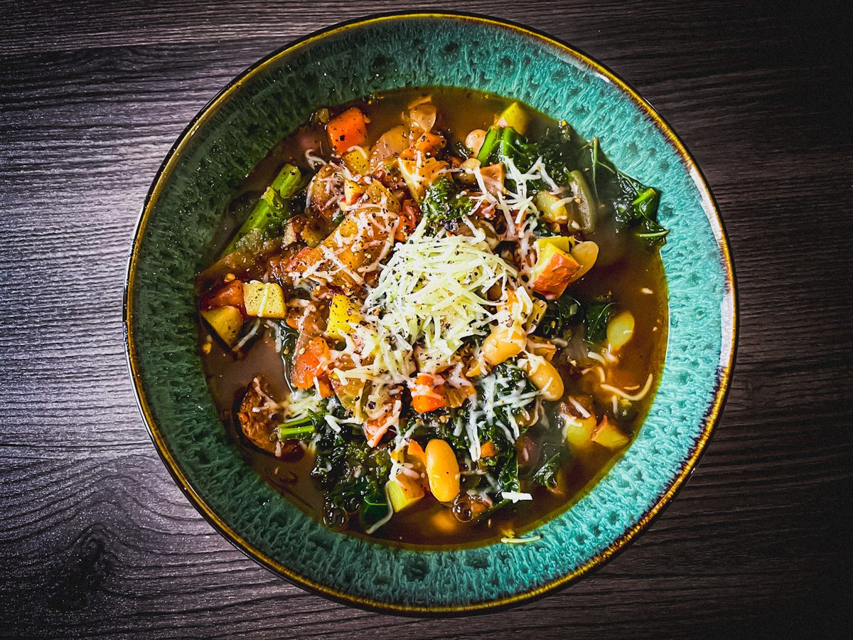 Teal glazed bowl of dark brothy soup with white beans, kale, carrot, zucchini and asparagus under shredded parmesan on a dark wood
