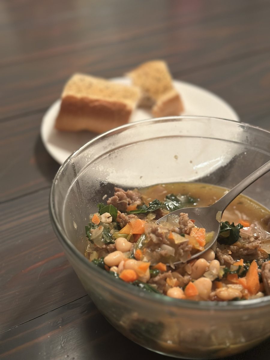 Glass bowl of white bean soup with crumbled sausage, kale and carrot with a spoon in it, a plate of bread chunks behind, on a dark