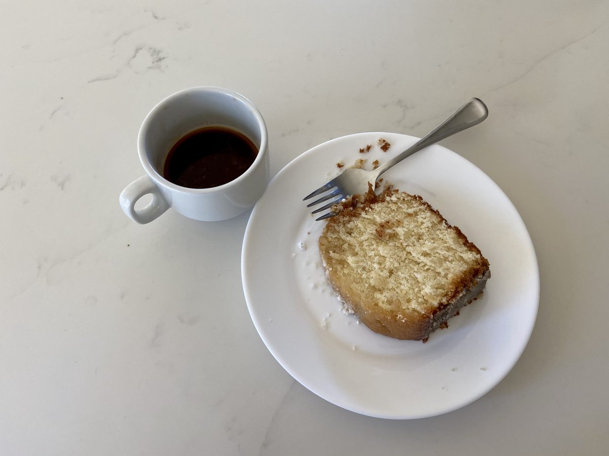 A slice of vanilla pound cake with a fork on a white plate beside a white cup of black coffee on a marble counter, crumbs scattere