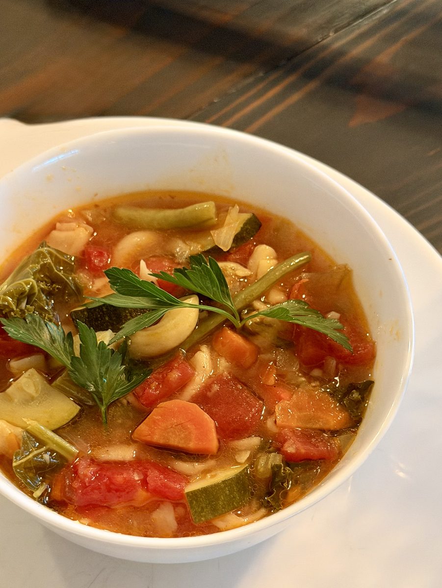 White bowl of chunky vegetable soup with zucchini, green beans, carrot, tomato, white beans and greens garnished with parsley, on