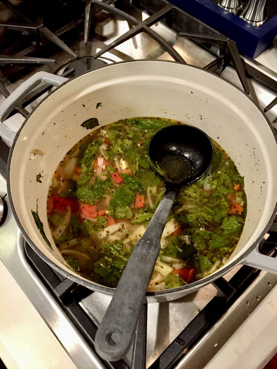 Overhead of a cream stockpot of kale and vegetable soup simmering on a gas range with a black ladle laid across it, cluttered dim