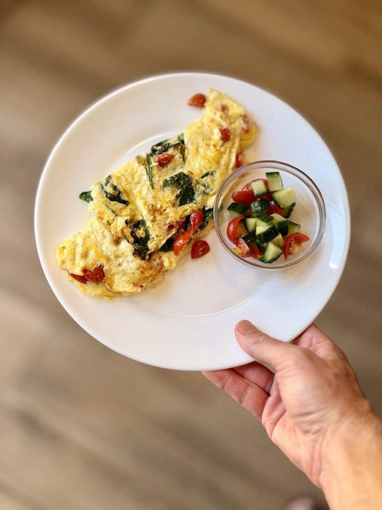 A plated veggie omelet with spinach and feta, held in one hand, with a small bowl of chopped tomato and cucumber