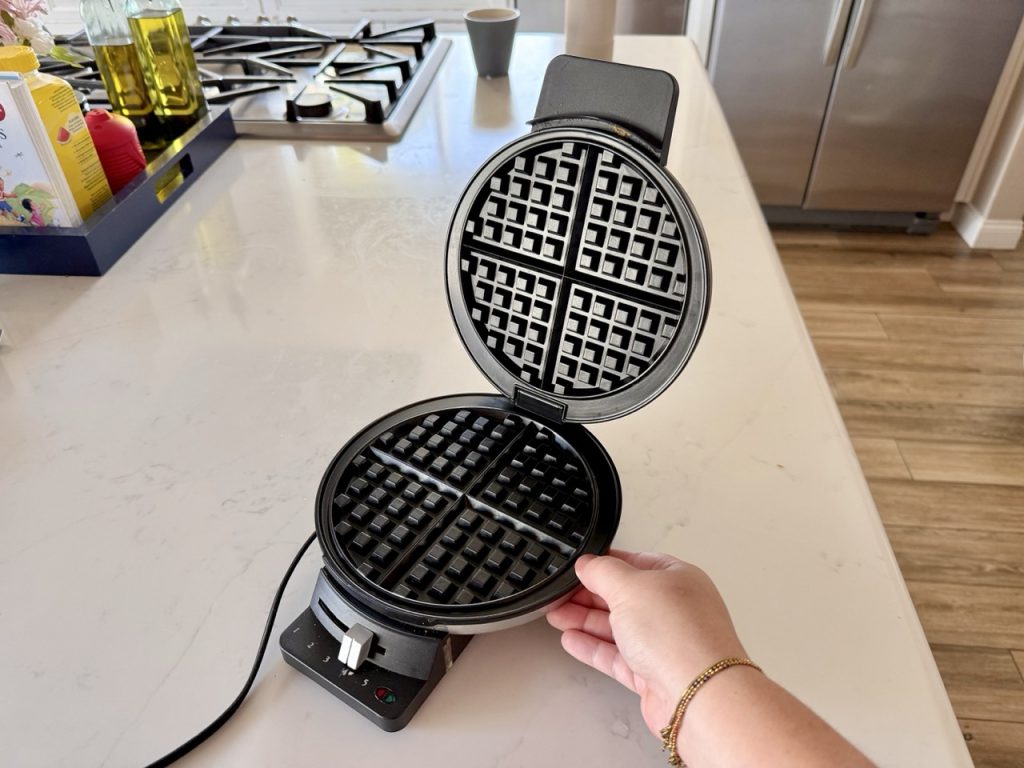 A hand opening a round waffle maker on a white kitchen counter, both plates empty