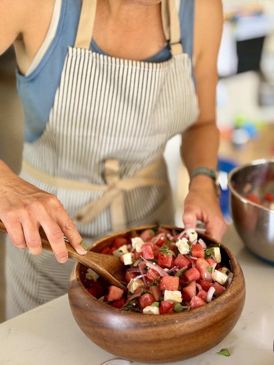 A woman in a striped apron (face out of frame) tossing cubed watermelon, feta, red onion and mint with a wooden spoon in a wooden