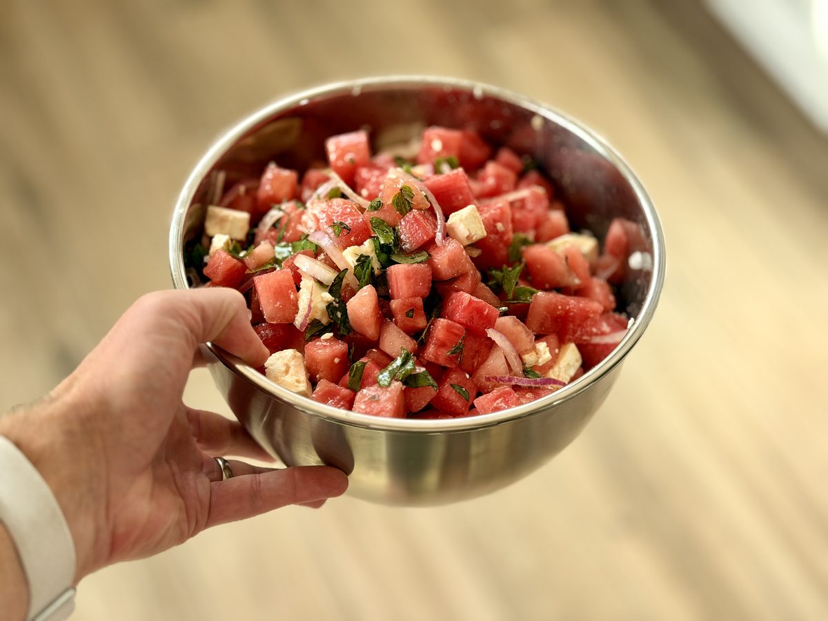 A hand holding out a stainless steel bowl piled with cubed watermelon, feta, red onion and mint, wooden floor blurred behind.