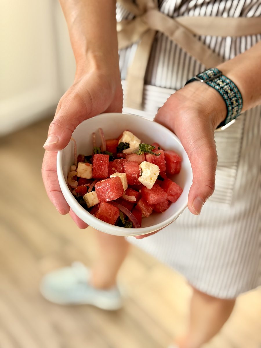 A person in a striped apron holding out a small white bowl of finished watermelon salad with cubed feta, slivered red onion and mi